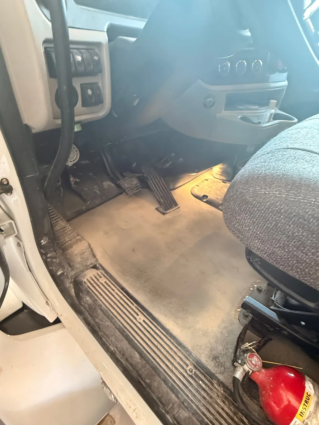 Cab floor of a semi truck with a sleeper before detailing, grey with dust around the pedals, a fire extinguisher beside the seat