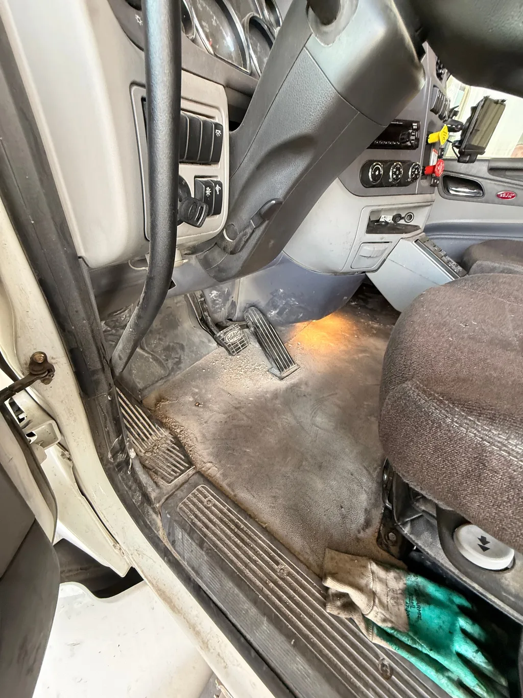 Driver side floor of a semi truck cab before detailing, caked with dried dirt around the pedals and door sill, work gloves by the seat